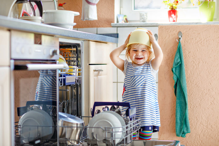 Happy toddler playing with a bowl on her head next to an open dishwasher in a kitchen.