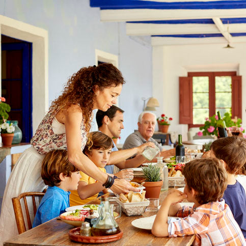  A multi-generational family gathers around a rustic table on a veranda, where a woman serves food to children. The setting is Mediterranean with blue and white decor.